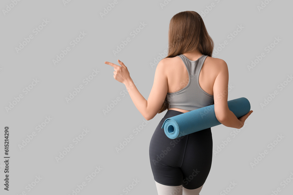 Sporty young woman with yoga mat pointing at something on grey background