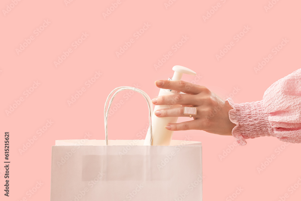 Hand of woman putting cosmetic product in shopping bag on pink background