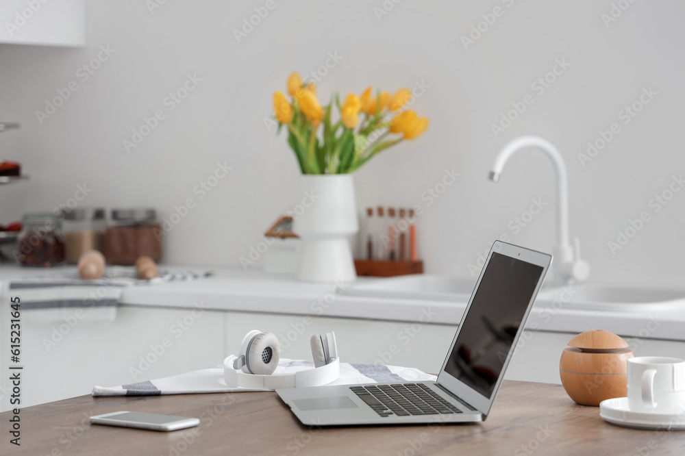 Modern laptop, headphones and mobile phone on wooden table in light kitchen