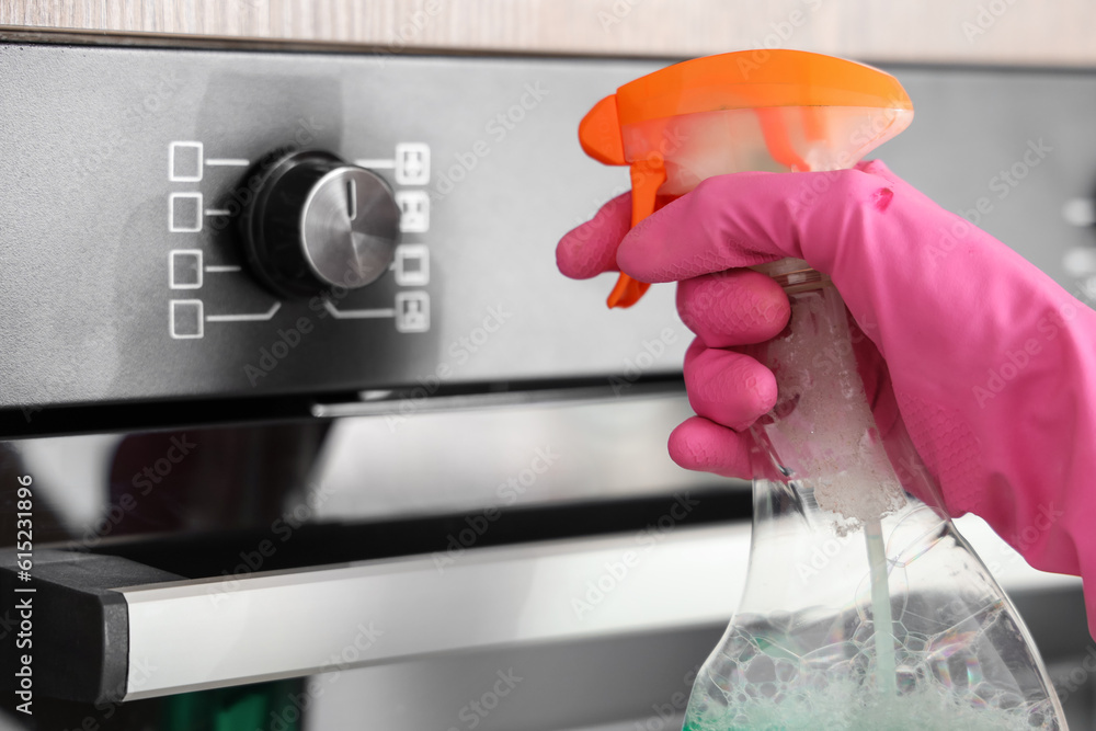 Woman in pink rubber glove cleaning electric oven with bottle of detergent, closeup