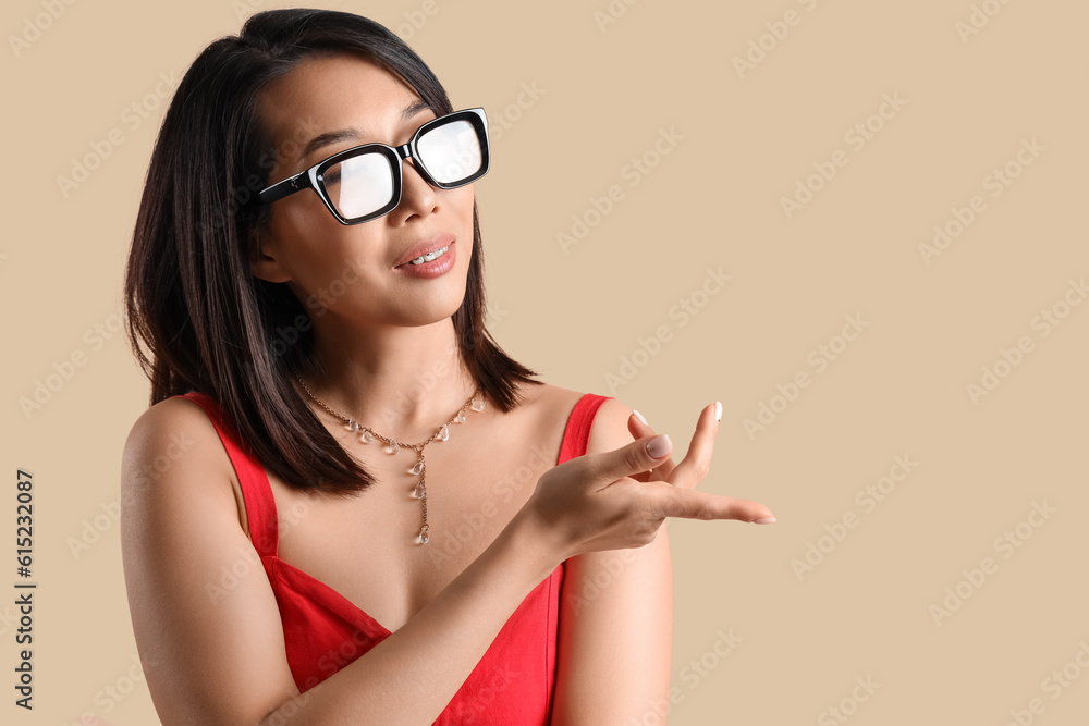 Beautiful Asian woman in stylish eyeglasses pointing at something on beige background