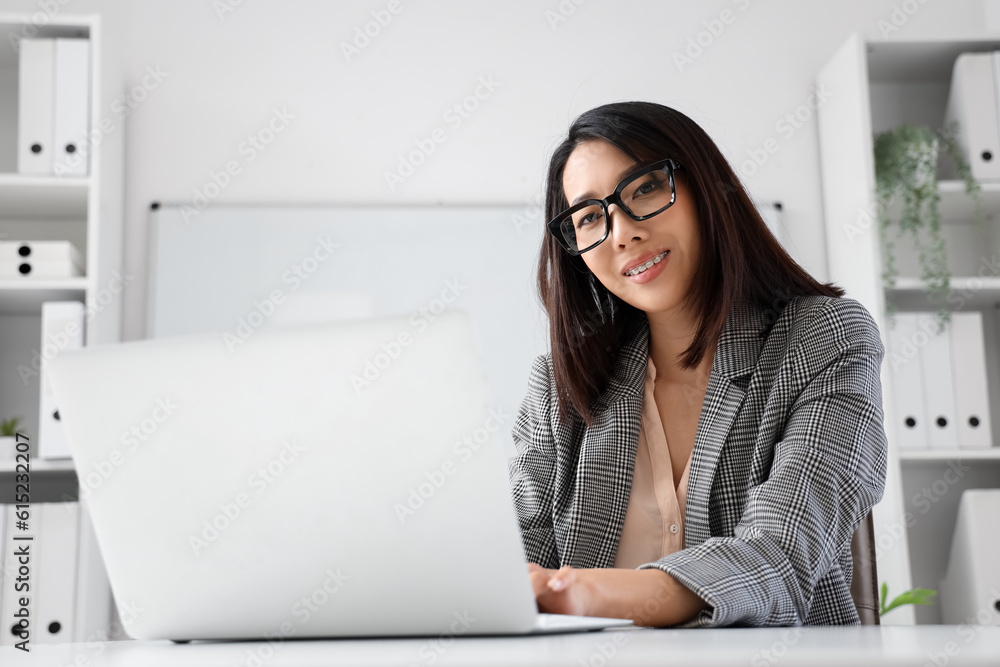 Beautiful Asian businesswoman in stylish eyeglasses working with laptop in office