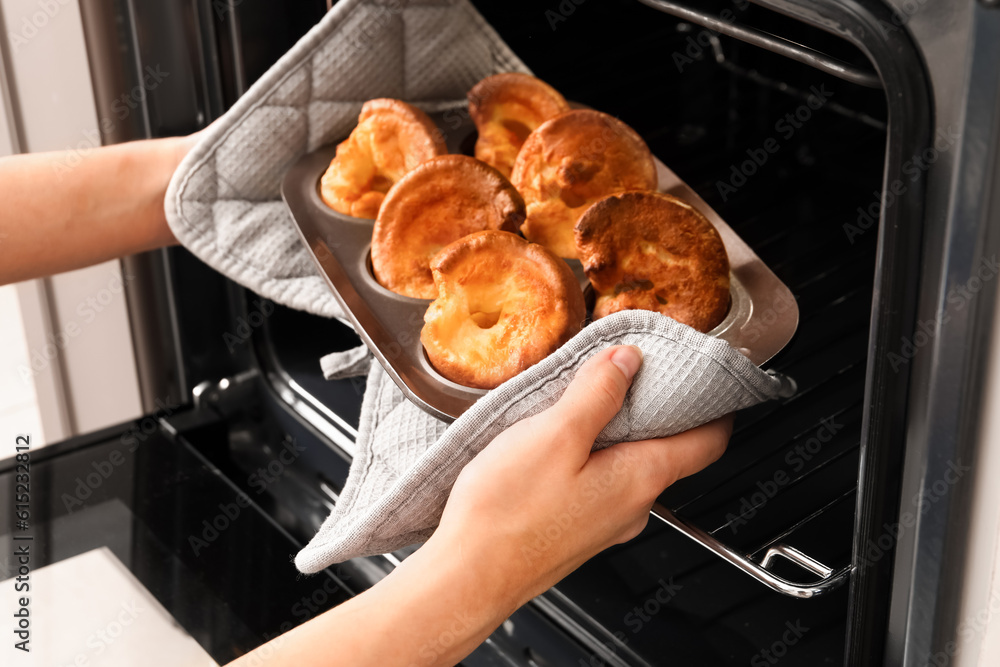 Woman taking baking tin with tasty Yorkshire pudding out from oven in kitchen