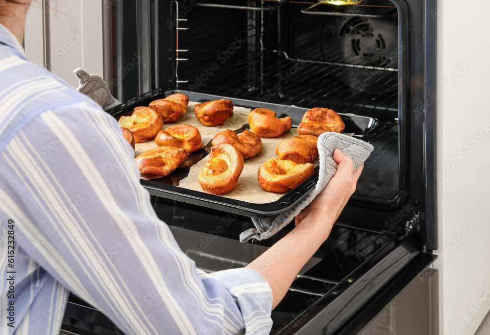 Woman taking baking tray with tasty Yorkshire pudding out from oven in kitchen