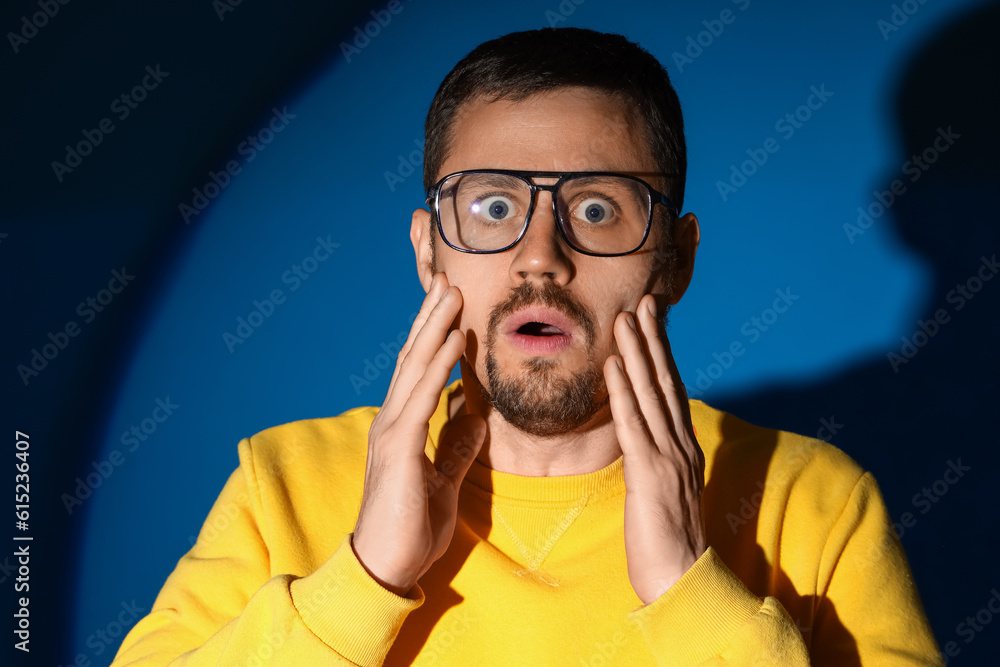 Shocked young man in stylish eyeglasses on dark blue background, closeup