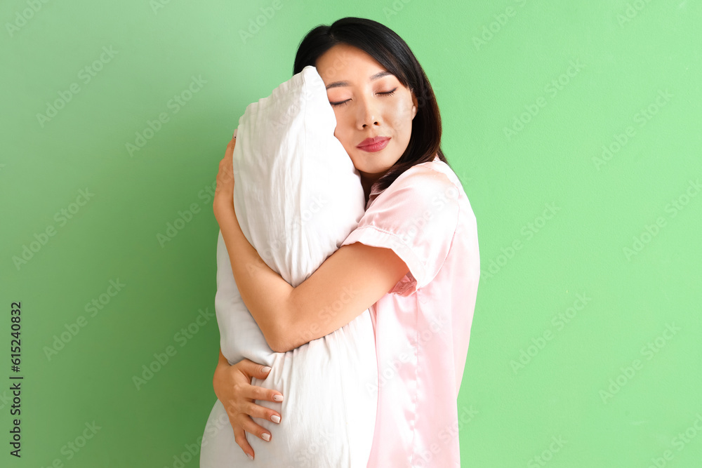 Beautiful Asian woman in pajamas with pillow on green background