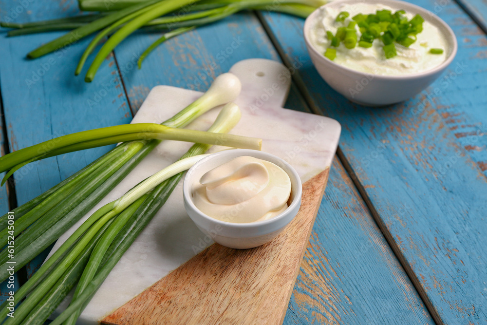 Board with bowls of tasty sour cream and green onion on blue wooden background