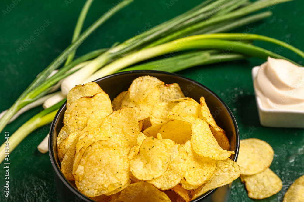 Bowl of tasty sour cream with scallion and potato chips on green background