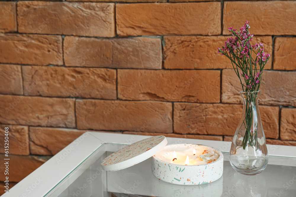 Beautiful flowers and burning candle on table near brick wall in room, closeup