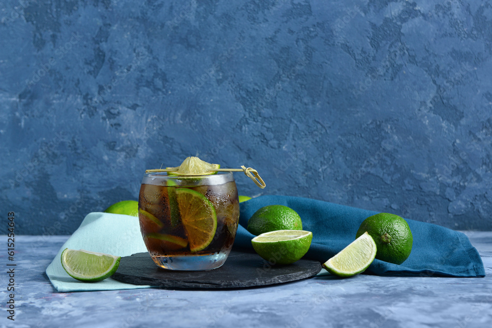 Board with glass of cold Cuba Libre cocktail and limes on blue background