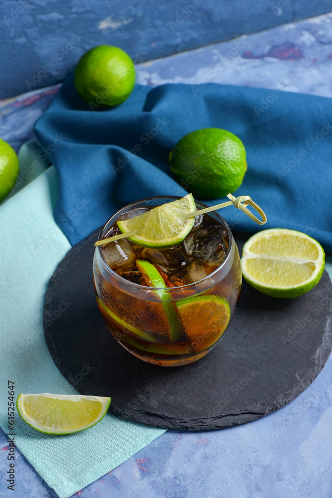 Board with glass of cold Cuba Libre cocktail and limes on blue background