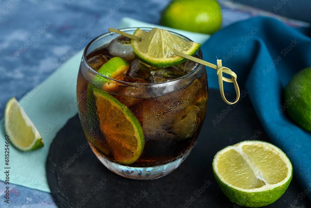 Board with glass of cold Cuba Libre cocktail and limes on blue background