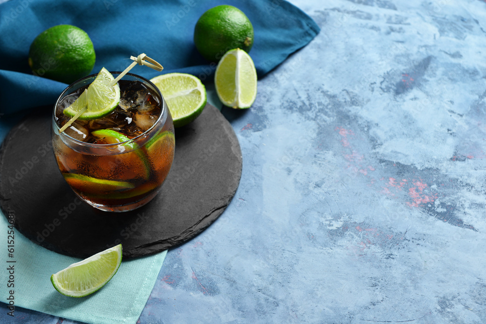 Board with glass of cold Cuba Libre cocktail and limes on blue background