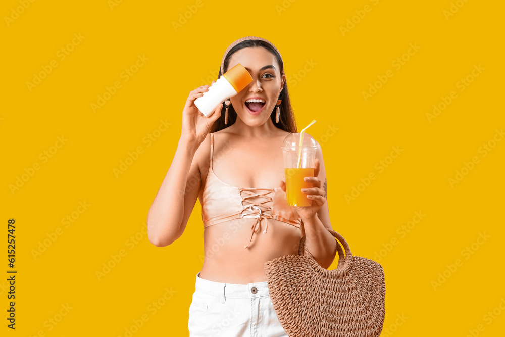 Young woman with sunscreen cream, juice and beach bag on yellow background