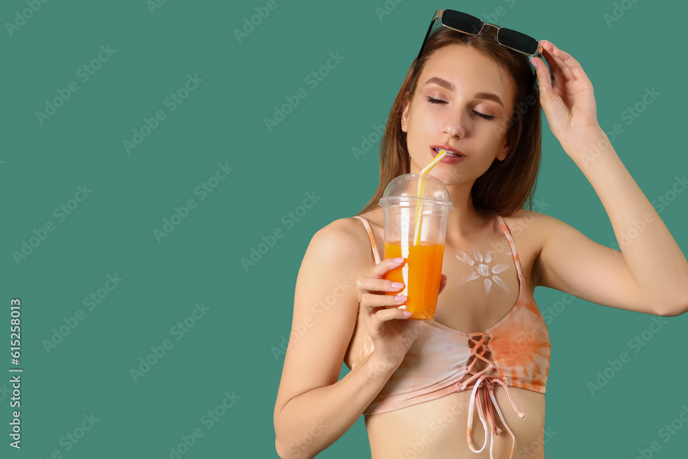 Young woman with sun made of sunscreen cream drinking juice on green background