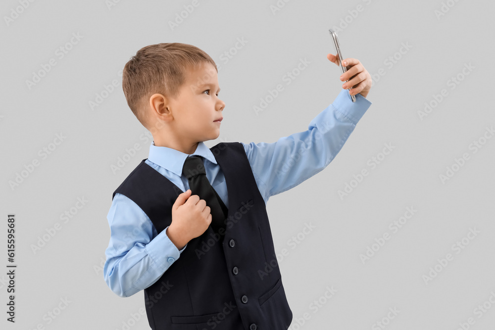 Cute little businessman with mobile phone taking selfie on grey background