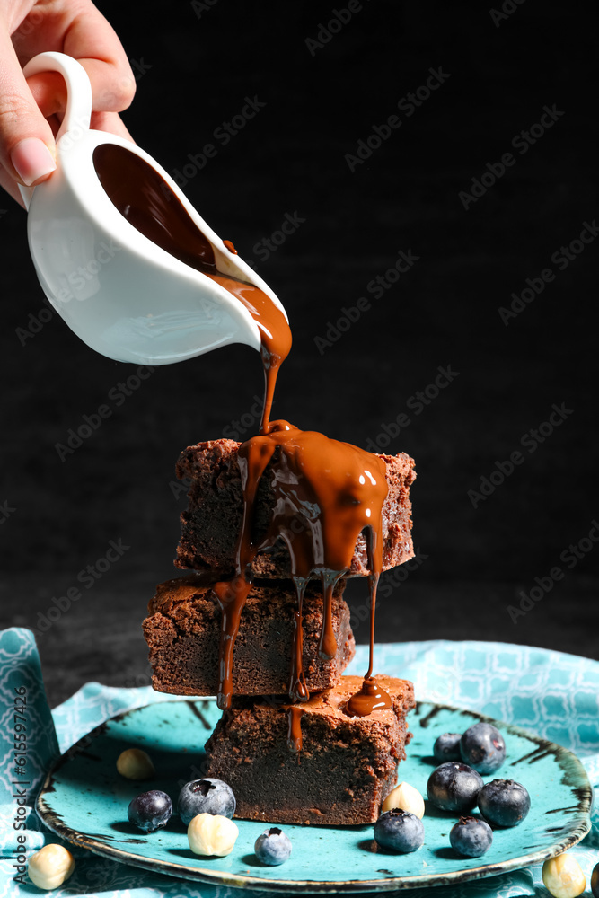 Woman pouring topping onto plate with pieces of tasty chocolate brownie on black background