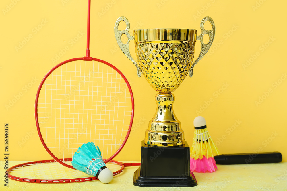 Gold cup with badminton rackets and shuttlecocks on yellow background