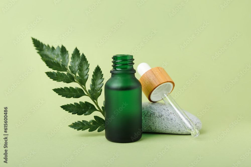 Open bottle of cosmetic oil with plant twig and stone on light green background