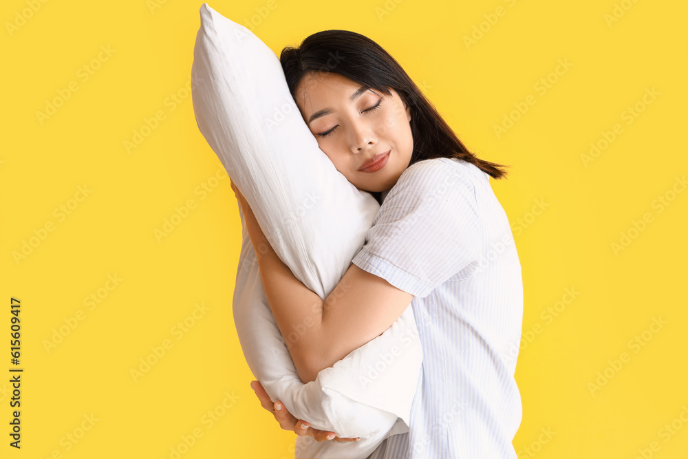 Beautiful Asian woman in pajamas with pillow on yellow background