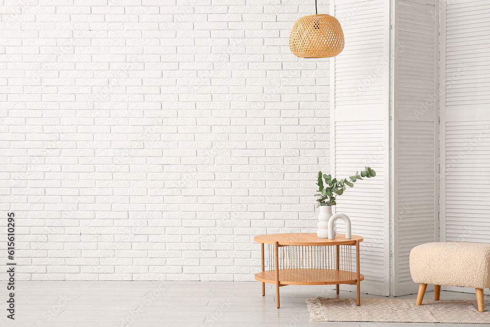 Wooden coffee table with eucalyptus branches in vase, folding screen and pouf near white brick wall