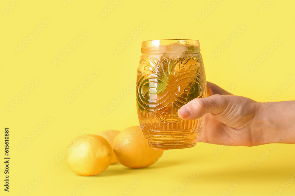 Female hand holding glass of ice tea with lemon and mint on yellow background