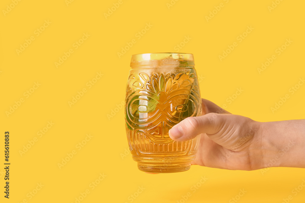 Female hand holding glass of ice tea with lemon and mint on yellow background