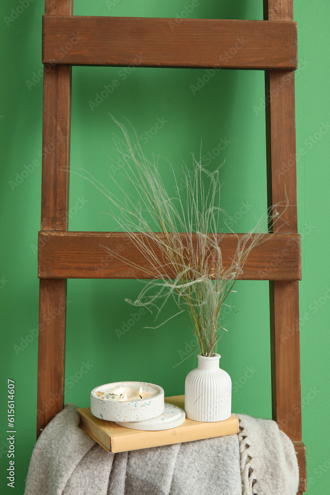 Burning candle, book and blanket on wooden ladder near green wall in room