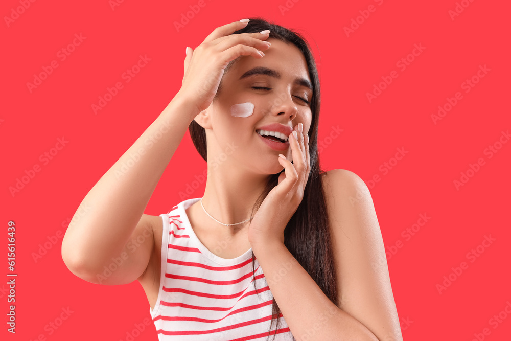 Young woman applying sunscreen cream onto her face against red background, closeup