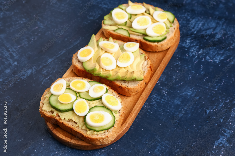 Tasty toasts with quail eggs, avocado and cucumber on dark blue background