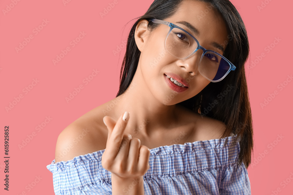 Beautiful Asian woman in eyeglasses making heart with her fingers on pink background, closeup