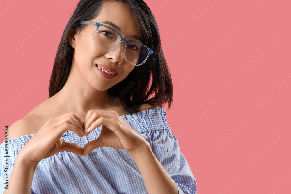 Beautiful Asian woman in eyeglasses making heart with her hands on pink background, closeup