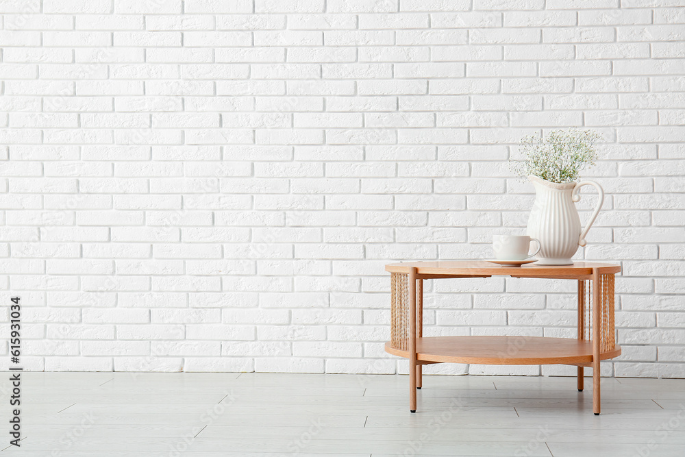 Vase with beautiful flowers and cup on wooden coffee table near white brick wall