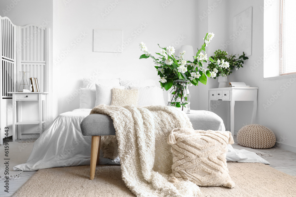 Vase with blooming jasmine flowers on bedside bench in interior of light bedroom