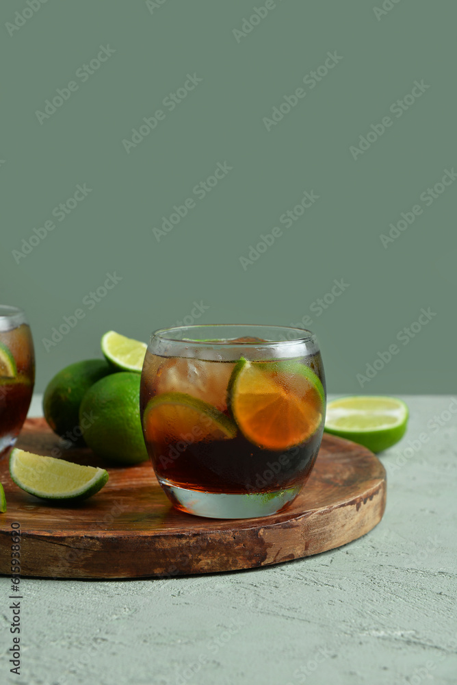 Board with glass of cold Cuba Libre cocktail and limes on grey background