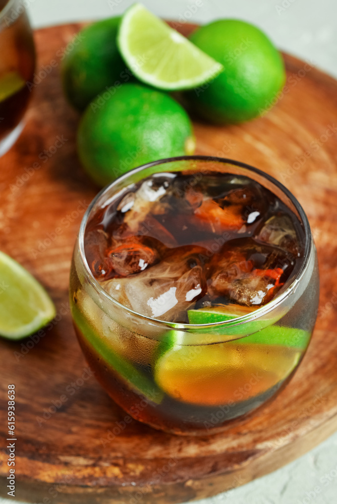 Board with glass of cold Cuba Libre cocktail and limes on grey background