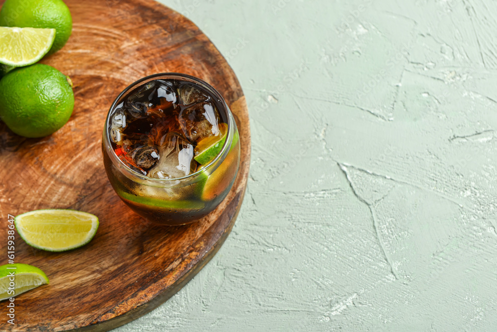 Board with glass of cold Cuba Libre cocktail and limes on grey background