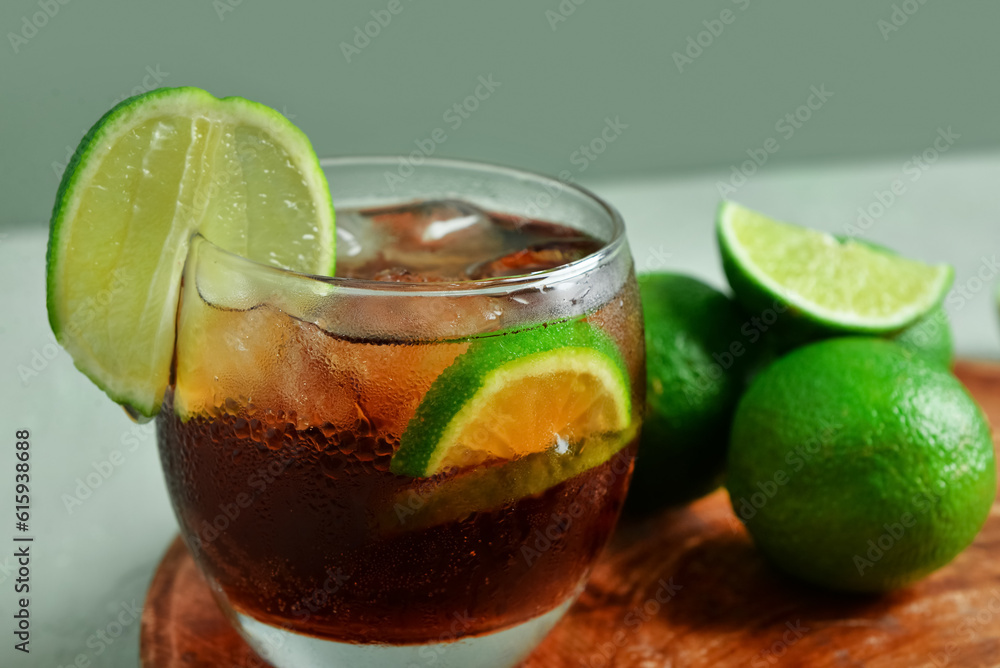 Board with glass of cold Cuba Libre cocktail and limes on grey background