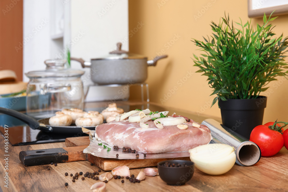 Aluminium foil roll with raw piece of meat, spices and vegetables on wooden table in kitchen