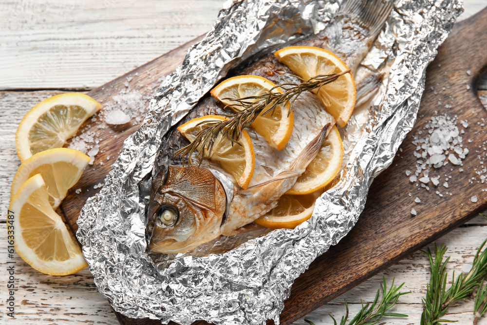 Aluminium foil with tasty baked fish, lemon slices and rosemary on light wooden background, closeup