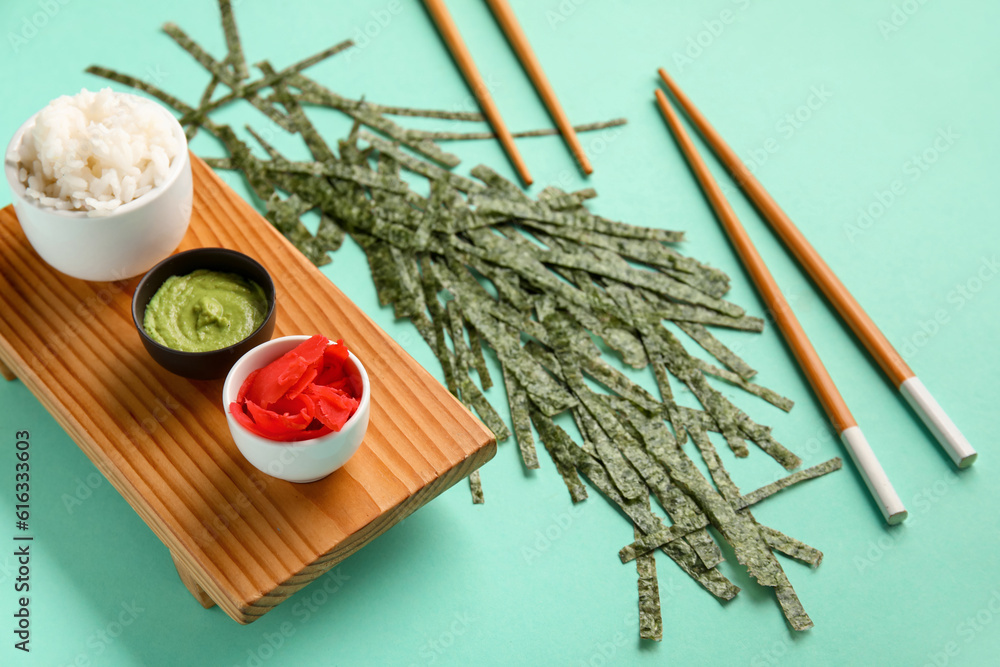 Wooden board with bowls of rice, ginger, wasabi and cut nori sheets on color background