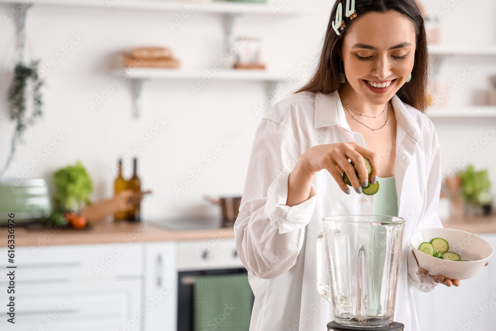 Young woman putting cucumber slices into blender in kitchen