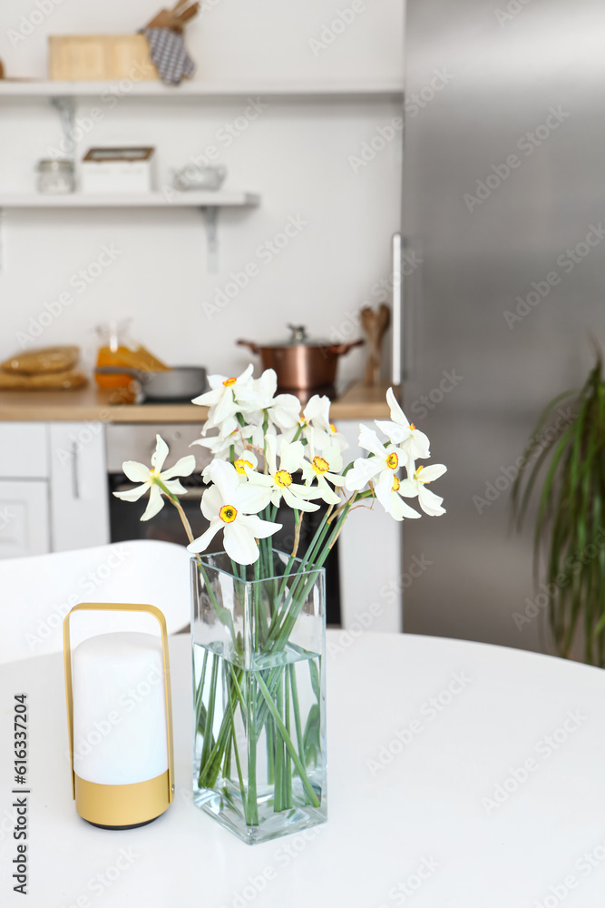 Vase with narcissus flowers and candle on table in interior of light kitchen, closeup