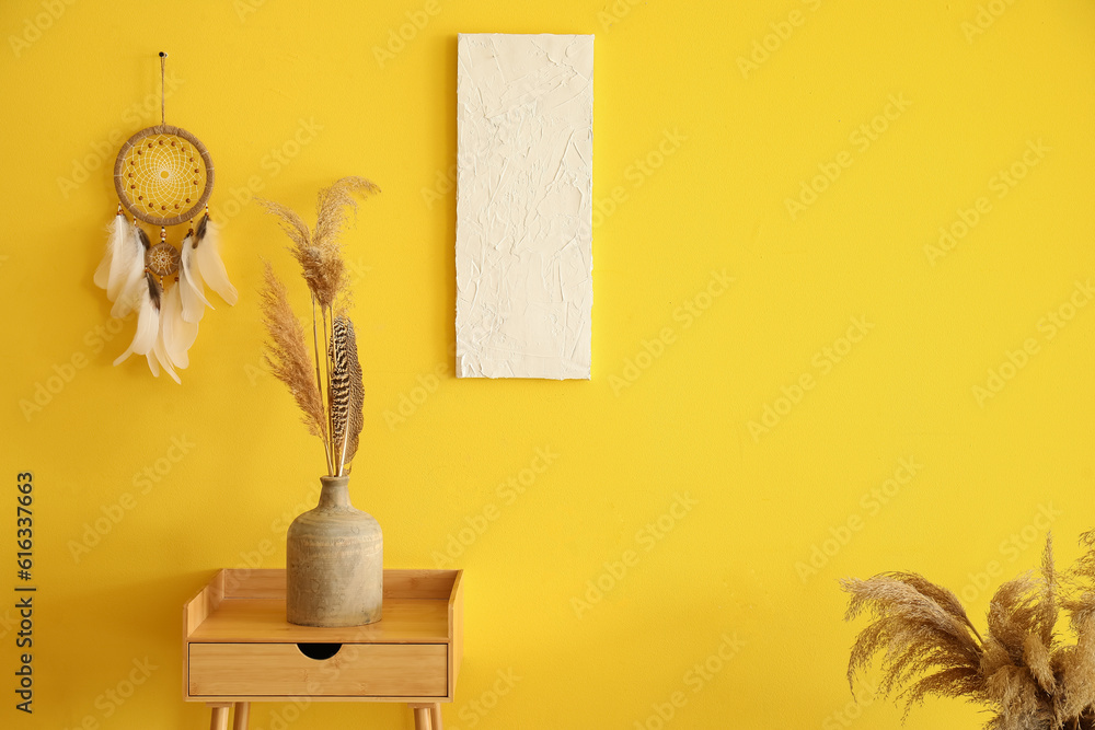 Table with vase of dried flowers and dream catcher hanging on yellow wall