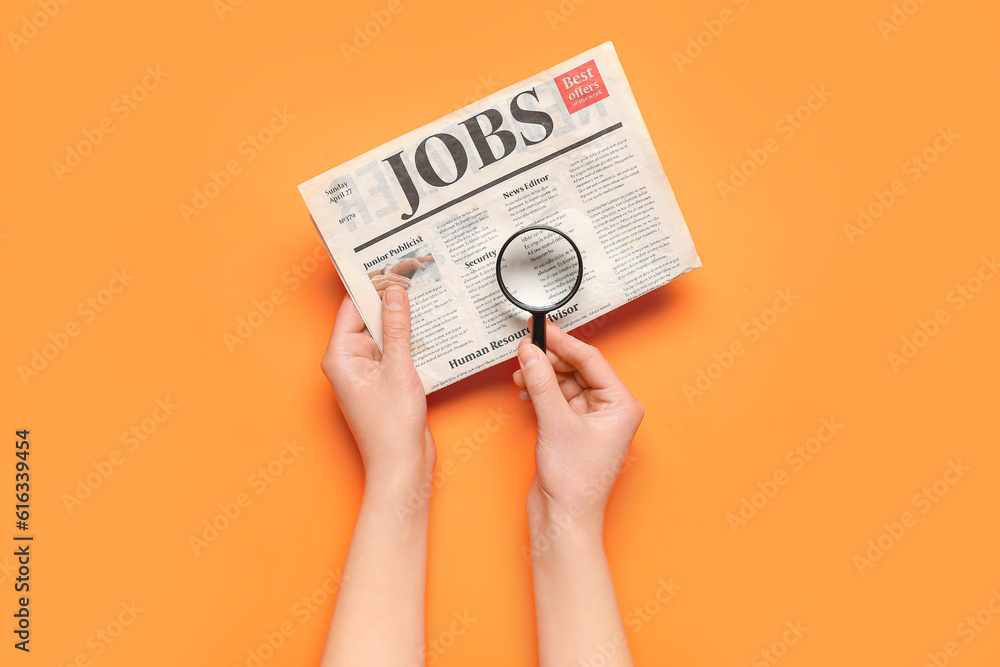 Female hands with mini magnifier and newspaper with headline JOBS on orange background. Search conce