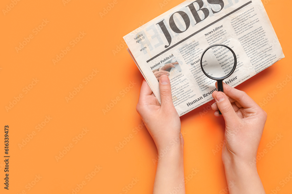 Female hands with mini magnifier and newspaper with headline JOBS on orange background. Search conce