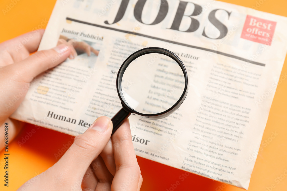 Female hands with mini magnifier and newspaper with headline JOBS on orange background. Search conce