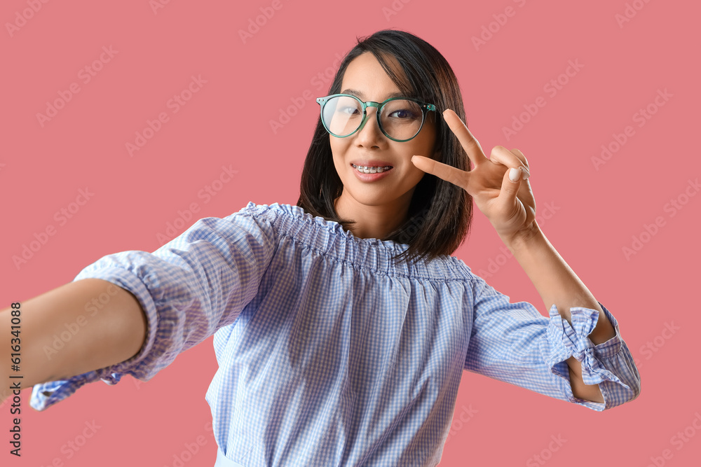 Beautiful Asian woman in stylish eyeglasses taking selfie on pink background