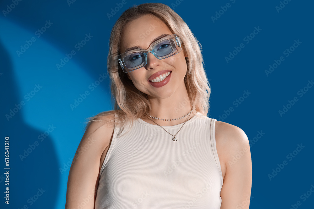 Young woman in stylish sunglasses on blue background, closeup