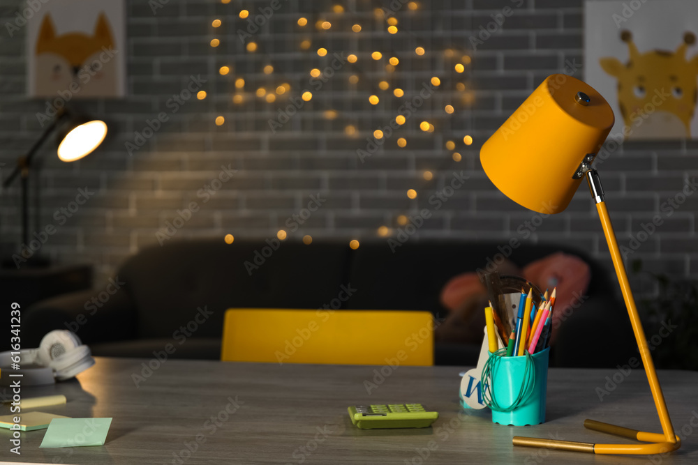 Table with school stationery and lamp in room at night